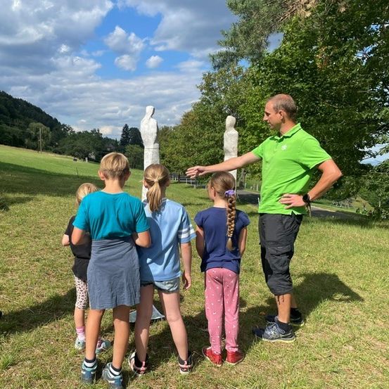 Ein Mann in einem grünen Shirt erklärt etwas einer Gruppe von Kindern auf einer Wiese mit Skulpturen im Hintergrund.