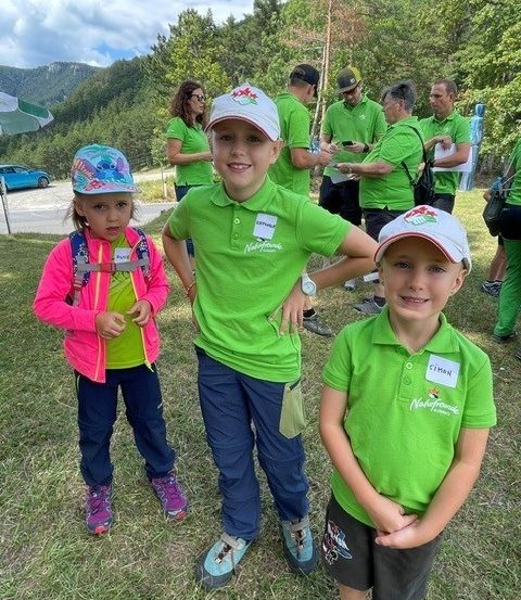 Zwei junge Kinder in grünen Hemden und Mützen stehen auf einem Rasenfeld mit Erwachsenen in grünen Hemden im Hintergrund.