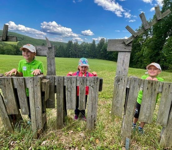 Drei Kinder, zwei Jungen und ein Mädchen, stehen hinter einem Holztor in einem Grasfeld mit blauem Himmel und Wolken im Hintergrund.