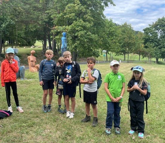 Eine Gruppe von Kindern in einem Park mit Rucksäcken, einige tragen Mützen, stehen in einer Reihe mit einem Grasfeld und Bäumen im Hintergrund.