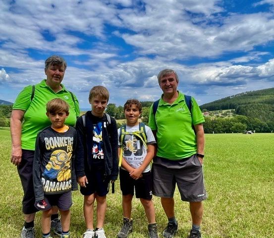 Eine Gruppe von Menschen, darunter zwei Erwachsene und drei Kinder, steht auf einer Wiese mit einem bewölkten Himmel im Hintergrund. Die Erwachsenen tragen grüne Hemden und Shorts, während die Kinder lässige Kleidung tragen. Sie alle lächeln und scheinen für ein Foto zu posieren.