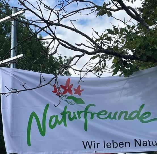 Ein weißes Banner mit grünem Text und roten Blumen steht 'Naturfreunde Wir leben Natur'. Es hängt von einem Ast.
