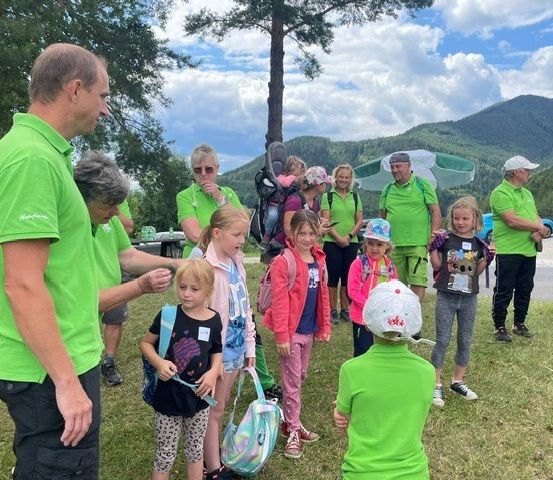 Eine Gruppe von Erwachsenen und Kindern in grünen Shirts steht auf einem Rasenfeld, wahrscheinlich in einem Park. Einige halten Taschen, andere schauen etwas an. Im Hintergrund befinden sich Berge, Bäume und ein Auto.