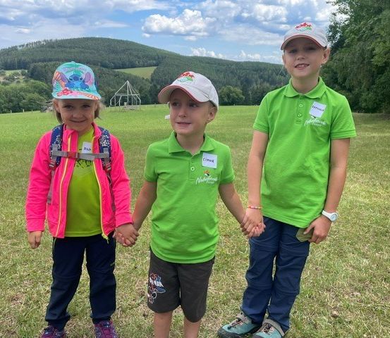 Drei Kinder, zwei Jungen und ein Mädchen, stehen in einem Grasfeld und halten Händchen. Sie sind in grüner und rosa Kleidung gekleidet und lächeln für die Kamera. Hinter ihnen ist ein malerischer Blick auf einen Hügel mit Bäumen.