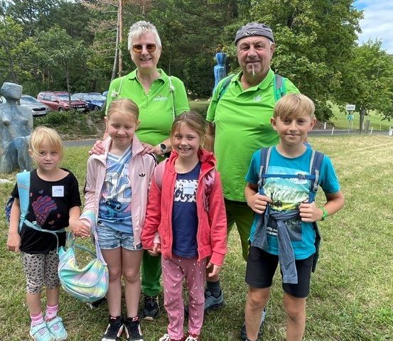 Eine Familie mit sechs Personen, darunter zwei Erwachsene und vier Kinder, steht in einem Park mit einem Grasfeld und Bäumen im Hintergrund.
