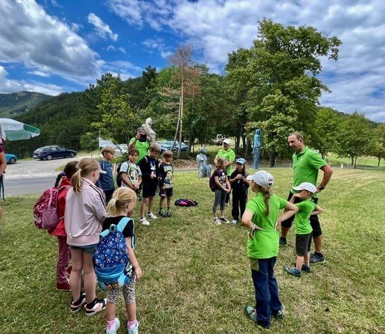 Eine Gruppe von Kindern und Erwachsenen in grünen Shirts versammelt sich auf einem Rasenfeld mit Bäumen und Bergen im Hintergrund. Einige tragen Rucksäcke.