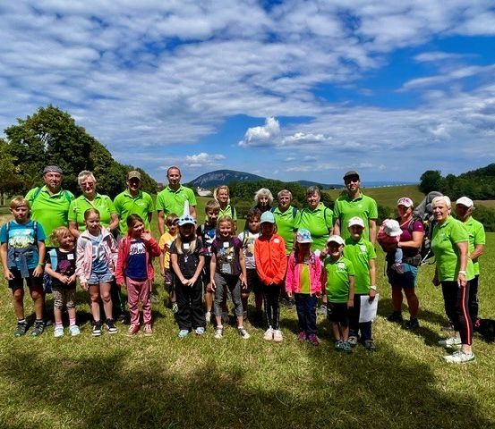 Eine Gruppe von Menschen, darunter Erwachsene und Kinder, steht auf einer Wiese und trägt grüne Hemden und Hüte. Sie lächeln und posieren für ein Foto. Im Hintergrund sind Bäume, ein Berg und ein blauer Himmel mit Wolken zu sehen.