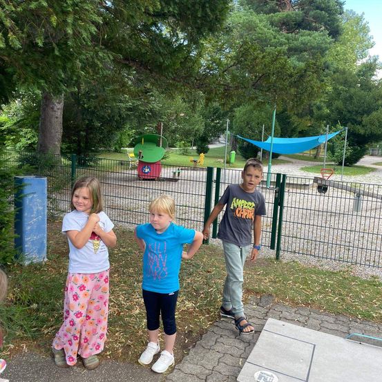 Drei Kinder stehen in einem Park, eine Mädchen in rosa Pyjamas, ein Junge in einem blauen T-Shirt und ein Junge in einem grauen T-Shirt, alle schauen zur Kamera. Dahinter befindet sich ein Spielplatz mit einem grünen Zaun und einem blauen Baldachin.