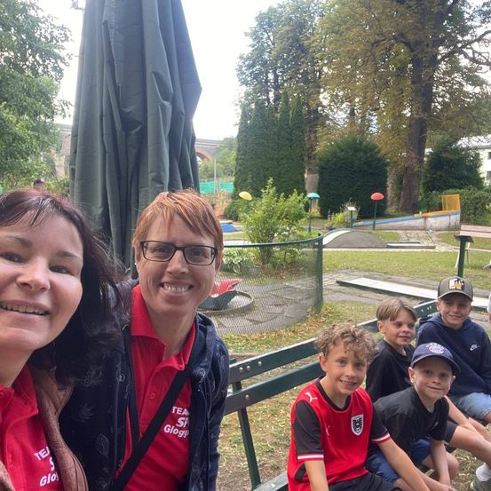 Eine Gruppe von Menschen, darunter zwei Frauen und vier Jungen, lächeln und posieren für ein Foto in einem Park. Die Frau auf der linken Seite trägt ein rotes Shirt mit dem Wort 'TEA' darauf. Die Frau in der Mitte trägt eine Brille und ein rotes Shirt. Die Jungen sitzen auf einer Bank. Hinter ihnen befindet sich ein Golfplatz mit Bäumen und einer Brücke.