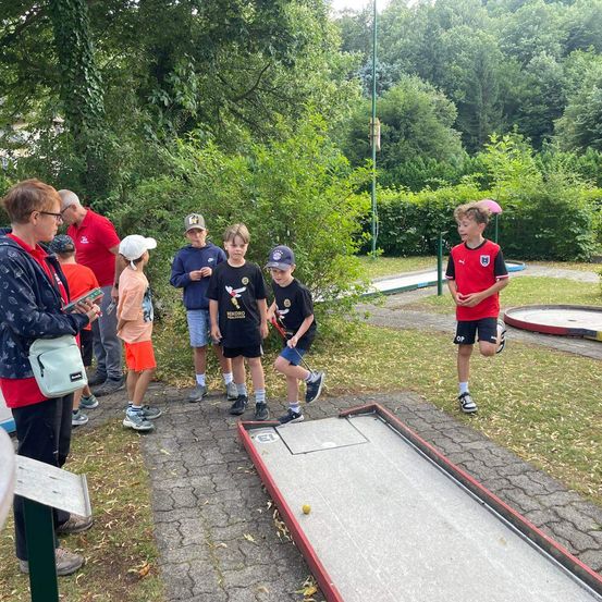 Eine Gruppe von Kindern und Erwachsenen versammelt sich um einen Minigolfplatz in einem Park. Ein Junge läuft auf das Loch zu.