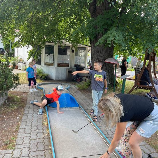Kinder spielen Minigolf im Park, ein Kind puttet, während ein anderes zuschaut. Bäume und ein kleines Gebäude sind im Hintergrund.