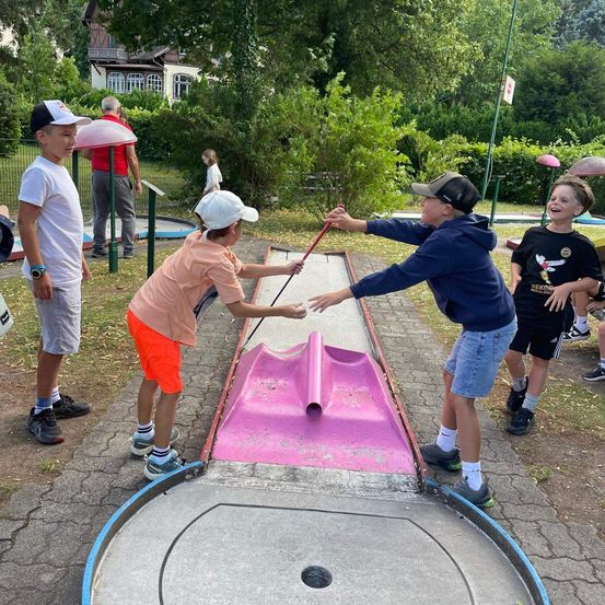 Vier Jungen spielen Minigolf in einem Park, wobei einer der Jungen kurz davor ist, den Ball zu schlagen. Bäume und ein Haus sind im Hintergrund.
