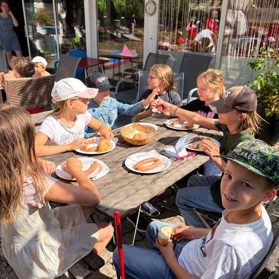 Eine Gruppe von Kindern sitzt um einen Tisch in einem Restaurant und isst Hot Dogs und lächelt.