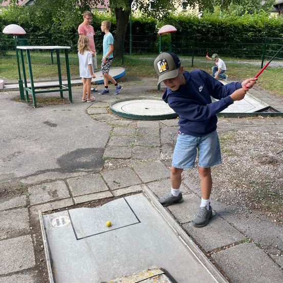 Ein Junge schwingt einen Golfschläger auf einem Übungsplatz, während andere zusehen und im Hintergrund spielen.