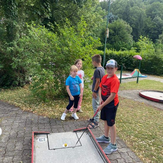 Vier Kinder spielen Minigolf in einem Park mit üppiger Grünfläche und blauem Himmel. Ein Junge ist kurz davor, den Ball zu schlagen.