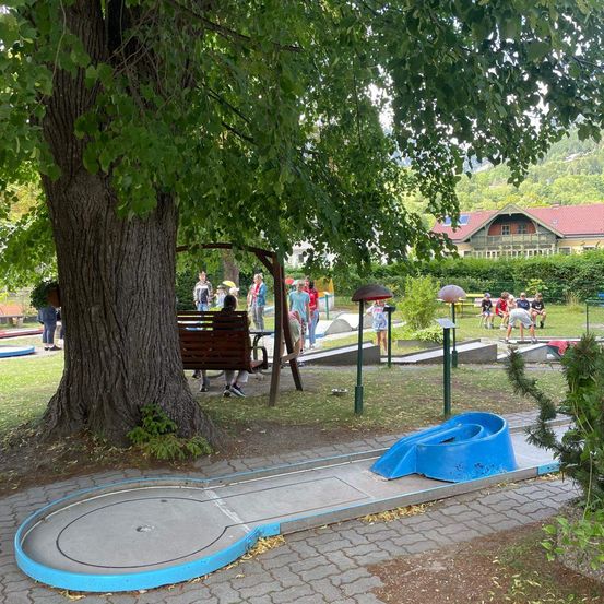 Ein Minigolfplatz mit einer blauen Rutsche unter einem Baum, spielende Personen und ein Haus im Hintergrund.
