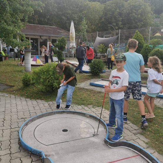 Eine Gruppe von Kindern spielt Minigolf in einem Außenbereich mit einem Haus und Bäumen im Hintergrund.