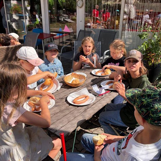 Eine Gruppe von Kindern sitzt um einen Holztisch herum und isst Hot Dogs. Einige von ihnen tragen Hüte. Ein Korb mit Hot Dogs steht auf dem Tisch. Hinter ihnen befinden sich Stühle und ein Tisch mit Tischdecke. Auf der rechten Seite steht ein Topf mit Pflanzen.