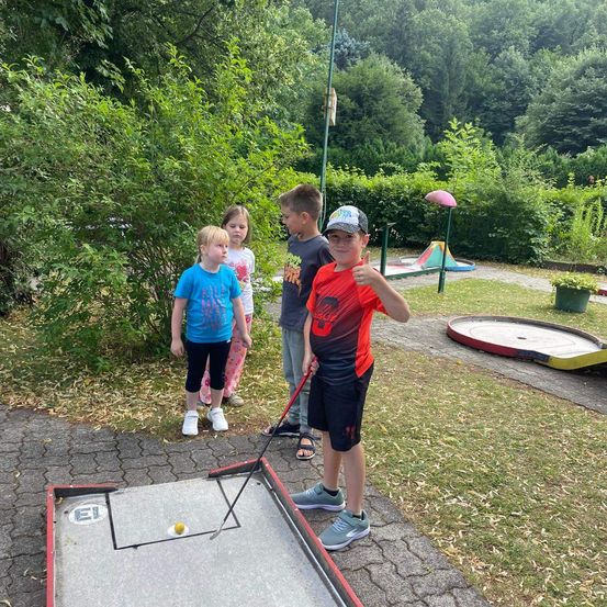 Vier Kinder spielen an einem sonnigen Tag Minigolf. Ein Junge ist dabei, den Ball zu schlagen, während die anderen zuschauen. Der Platz ist von Grün umgeben.