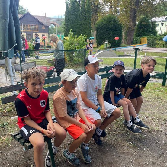 Fünf Jungen sitzen auf einer Parkbank. Ein Junge trägt ein rotes Fußballtrikot. Dahinter befindet sich ein Zaun mit einem Spielplatz.