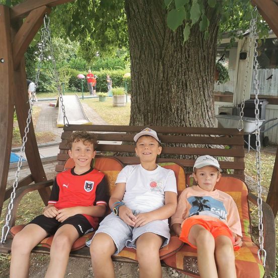 Drei junge Jungen sitzen auf einer Schaukel unter einem großen Baum in einem Park. Der Junge auf der linken Seite trägt ein rotes Fußballtrikot. Der Junge in der Mitte trägt ein weißes Hemd und eine Kappe. Der Junge auf der rechten Seite trägt ein orangefarbenes Hemd.