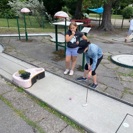 Zwei Mädchen spielen Minigolf an einem sonnigen Tag. Ein Mädchen beugt sich vor, um zu zielen, während das andere zusieht.