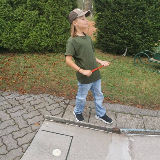 Ein junger Junge in einem grünen T-Shirt und Jeans steht auf einem Betonweg, hält einen Golfschläger und schaut nach oben. Hinter ihm befinden sich Büsche und ein grüner Schubkarren.