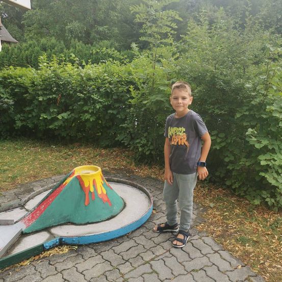 Ein junger Junge steht neben einem Minigolfplatz mit einem vulkanartigen Design, trägt ein graues T-Shirt mit einem farbenfrohen Grafik und einer Uhr.