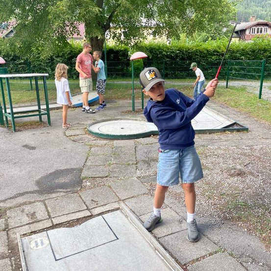 Ein junger Junge schwingt einen Golfschläger auf einem Minigolfplatz, während einige Leute im Hintergrund zusehen.