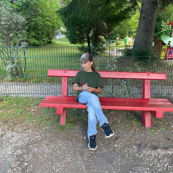 Ein junger Junge sitzt auf einer roten Bank im Park, hält ein Notizbuch und einen Stift in der Hand und trägt ein grünes Hemd, Jeans und Turnschuhe.