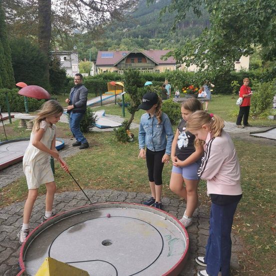 Eine Gruppe von Kindern und Erwachsenen spielt Minigolf in einem Park mit Bäumen und Bergen im Hintergrund.