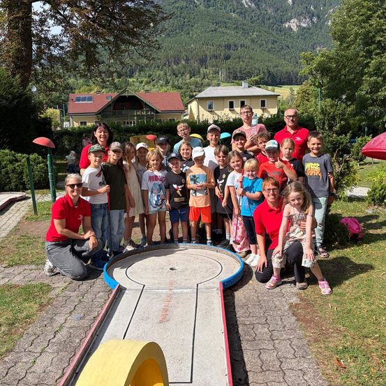 Eine Gruppe von Kindern und Erwachsenen posiert für ein Foto vor einem Minigolfplatz mit Bergen im Hintergrund.
