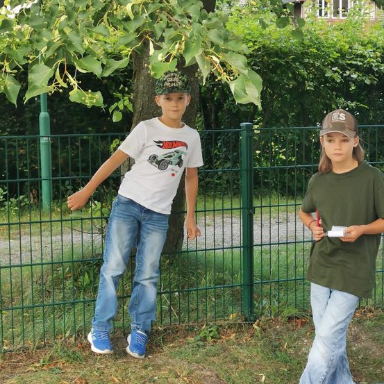 Zwei Kinder stehen neben einem grünen Zaun, eines trägt ein Hot Wheels T-Shirt und das andere hält eine Karte.