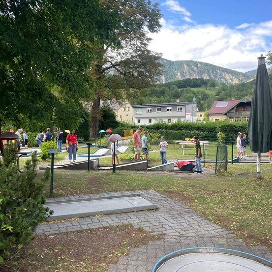 Eine Gruppe von Menschen spielt Minigolf an einem sonnigen Tag, mit Bergen im Hintergrund und Häusern in der Nähe.