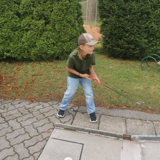Ein junger Junge in einem grünen T-Shirt und Jeans übt Golf auf einem Betonweg, umgeben von Hecken und Gras.