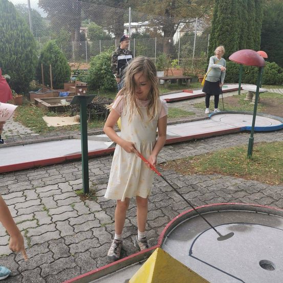 Ein kleines Mädchen in einem Blumenkleid spielt Minigolf und hält einen Schläger in der Hand, während eine Frau und ein Junge im Hintergrund stehen.