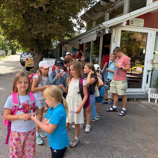 Eine Gruppe von Kindern und Erwachsenen steht in einer Schlange vor einem Gebäude mit Glastüren, wahrscheinlich warten sie darauf, einzutreten. Einige Kinder tragen Rucksäcke und Hüte. Ein Mann in einem rosa T-Shirt steht in der Nähe des Eingangs.