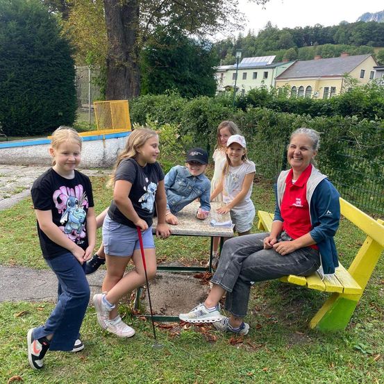Eine Gruppe von Kindern und eine erwachsene Frau stehen um ein kleines Golf-Putt-Grün in einem Park. Die Frau sitzt auf einer gelben Bank und lächelt.