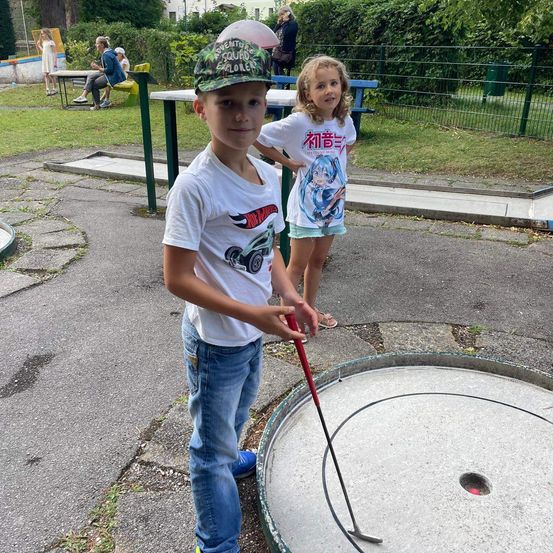 Ein junger Junge und ein Mädchen spielen Minigolf in einem Park. Der Junge hält einen Golfschläger und steht vor einem Loch. Das Mädchen steht hinter ihm.