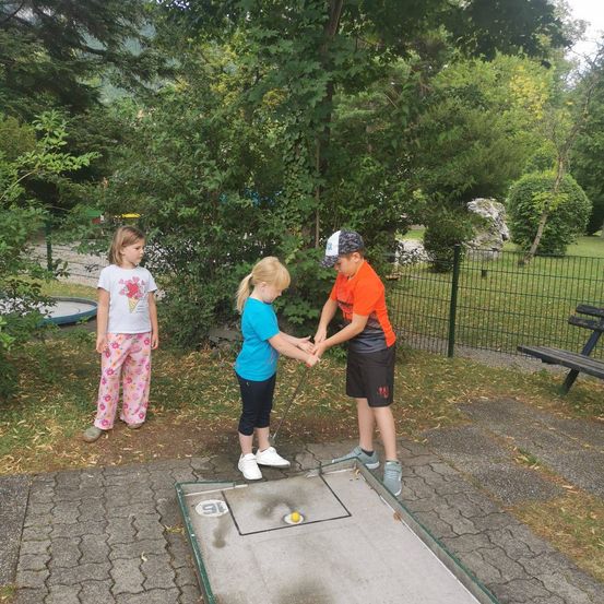 Drei Kinder spielen Golf auf einem kleinen Platz. Ein Mädchen in einem blauen Shirt und ein Junge in einem orangefarbenen Shirt halten sich an den Händen und schlagen einen Golfball. Ein weiteres Mädchen in rosa Hosen steht hinter ihnen.