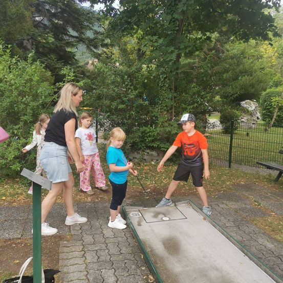 Eine Frau und vier Kinder spielen Minigolf in einem Park. Die Frau beobachtet den Jungen beim Putten.