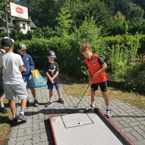 Fünf Kinder spielen an einem sonnigen Tag Minigolf. Ein Junge ist dabei, den Ball zu schlagen.