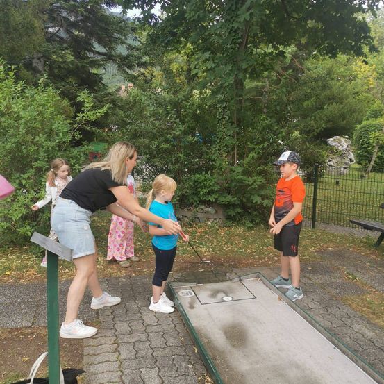 Eine Frau hilft einem kleinen Mädchen mit einem Golfschläger auf einem Minigolfplatz, während ein Junge in der Nähe zuschaut.