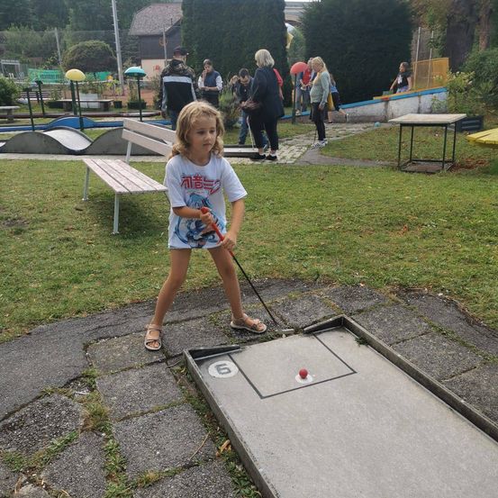 Ein kleines Mädchen spielt Minigolf in einem Park. Sie steht auf einer Betonplattform mit einem Golfschläger und einem roten Ball. Hinter ihr gehen und stehen Leute, und in der Nähe ist eine Bank.