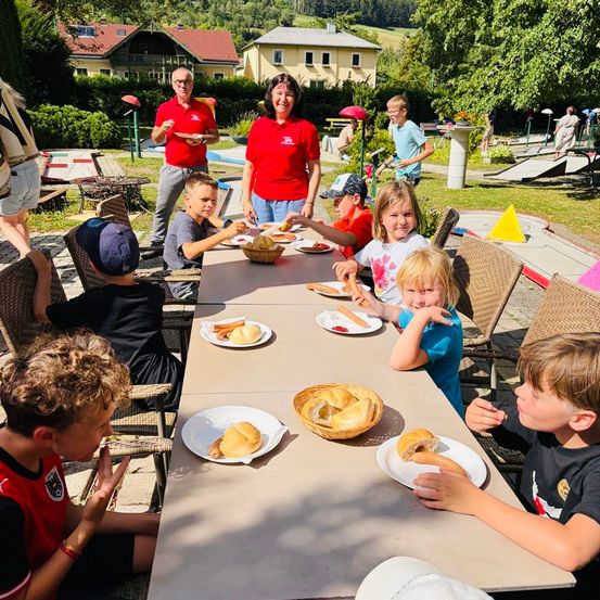 Eine Gruppe von Kindern und Erwachsenen sitzt um einen Tisch in einer Outdoor-Umgebung, möglicherweise einem Park, und genießt eine Mahlzeit. Die Kinder essen Hot Dogs und Sandwiches. Eine Frau in einem roten Shirt steht hinter dem Tisch, und ein Mann in einem roten Shirt steht in der Nähe. Im Hintergrund befinden sich Häuser und ein Golfplatz.