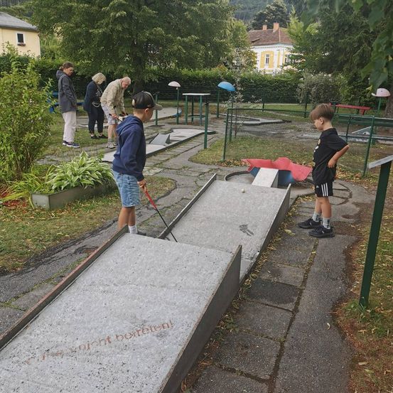Eine Gruppe von Menschen spielt Minigolf in einem Park. Ein Junge in einer blauen Kapuzenjacke ist dabei, den Ball zu schlagen.