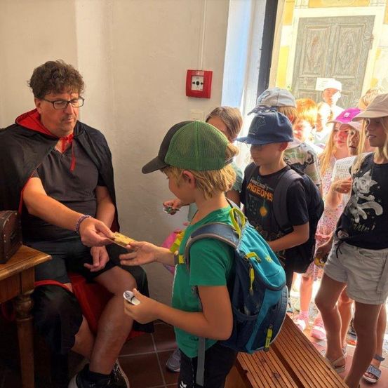 Ein Mann in einem Kostüm gibt einem Jungen in einem grünen Hut eine goldene Münze, umgeben von anderen Kindern in einem Raum mit einer Holzbank und einem Feuerlöscher an der Wand.