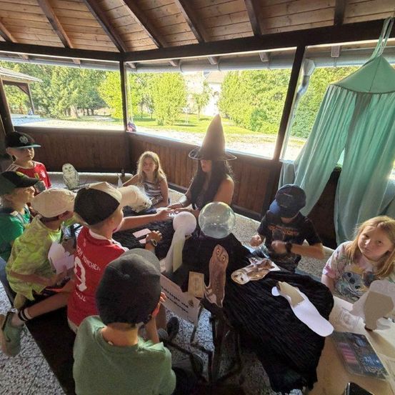 Eine Gruppe von Kindern in einem Pavillon, einige mit Hüten, sind in einer Bastelaktivität mit einer als Hexe gekleideten Frau engagiert. Die Umgebung ist im Freien mit Bäumen, die durch die Fenster sichtbar sind.
