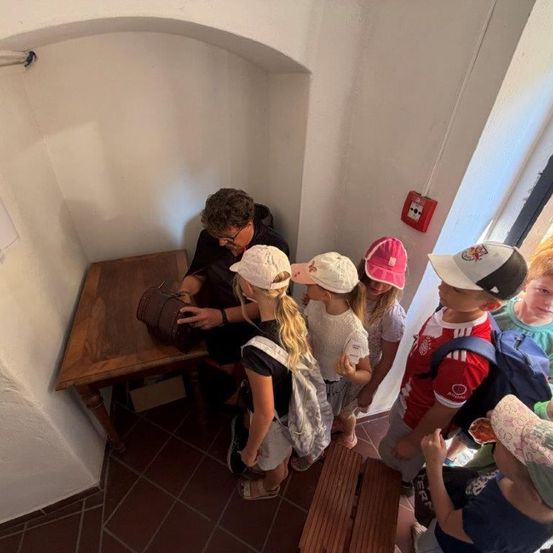 Ein Mann zeigt einer Gruppe von Kindern einen braunen Beutel in einem Raum mit gefliesten Böden und weißen Wänden.