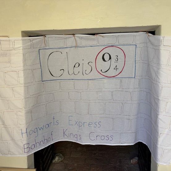 Ein weißer Banner mit einem Ziegelwandhintergrund zeigt den Text 'Gleis 9 3/4' und 'Hogwarts Express Bahnhof Kings Cross' in Blau. Der Banner hängt an einer Wand.